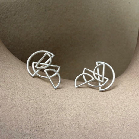 A pair of silver geometric stud earrings displayed on a textured beige surface. By contemporary jewellery designer, Sarah Straussberg
