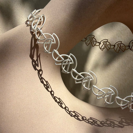A sterling silver necklace with interlocking geometric links, draped on a ceramic sculpture. The necklace is shown in warm sunlight with a distinct shadow cast on the form, and a darker, out-of-focus version of the same necklace is visible in the background. By contemporary jewellery designer, Sarah Straussberg
