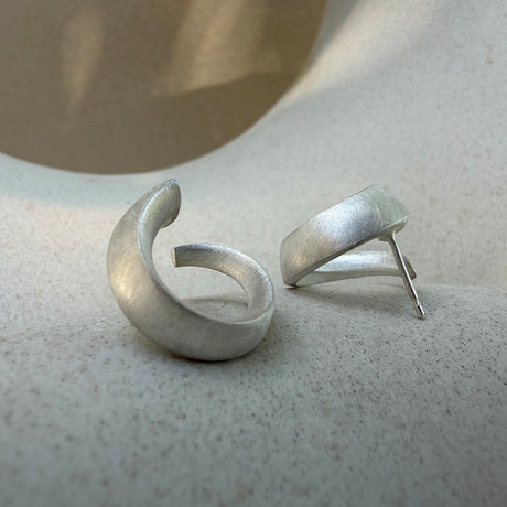 A pair of textured sterling silver sculptural loop earrings, one shown from the front and the other from the side to display the post, resting on a light beige stone surface. By contemporary jewellery designer, Sarah Straussberg