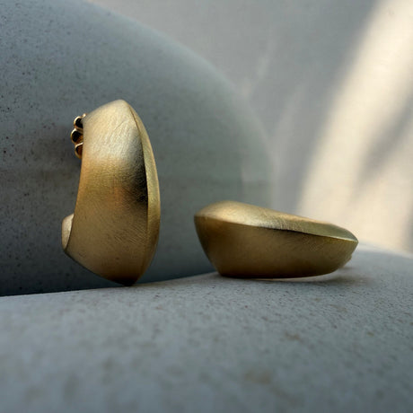 Two matte gold dome-shaped earrings displayed on a textured grey stone background. By contemporary jewellery designer, Sarah Straussberg