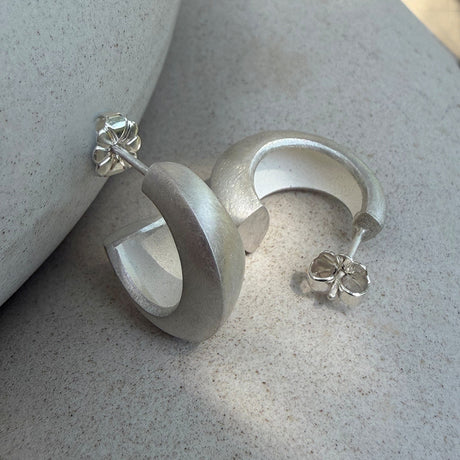 A pair of matte silver chunky hoop earrings displayed on a textured light grey stone surface. By contemporary jewellery designer, Sarah Straussberg